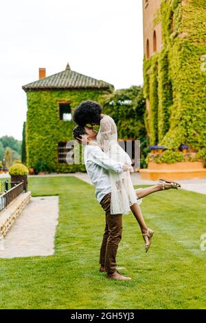 Vue latérale de l'homme levant la femme noire en se tenant sur la prairie dans le jardin contre le bâtiment avec la lierre sur le mur Banque D'Images