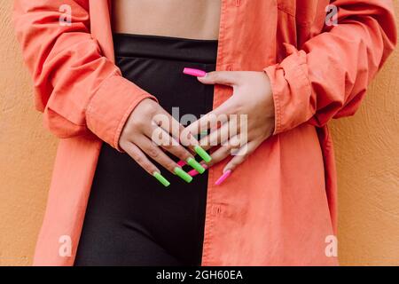 Rognez une femme méconnaissable avec de longs ongles colorés en portant une chemise brillante près du mur orange Banque D'Images