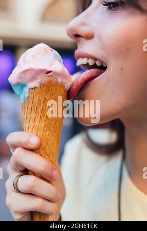Croquer une jeune femme souriante et déguster une glace savoureuse dans un cône à gaufres tout en regardant loin en ville sur un fond flou Banque D'Images