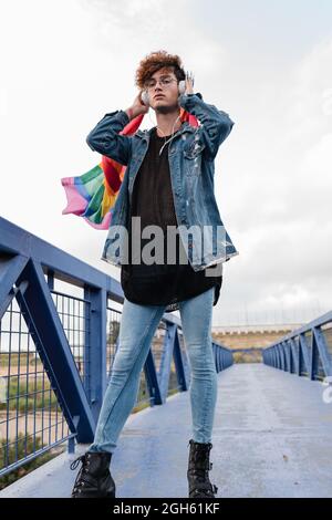 Homosexuel élégant homme avec drapeau LGBT coloré debout sur le pont et écouter de la musique dans le casque tout en regardant l'appareil photo Banque D'Images