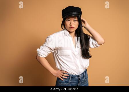 Vue avant du modèle asiatique féminin tendance en chemise blanche et Jean touchant la casquette sur fond beige et regardant l'appareil photo Banque D'Images
