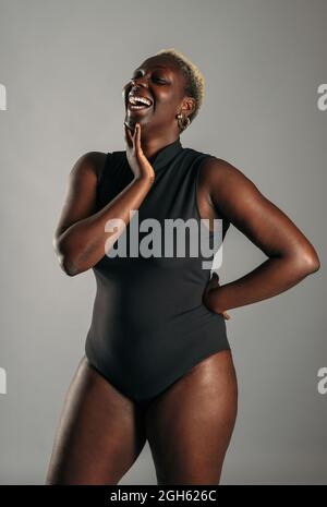 Gaie afro-américaine curvy femme en body noir debout sur fond gris en studio et riant Banque D'Images