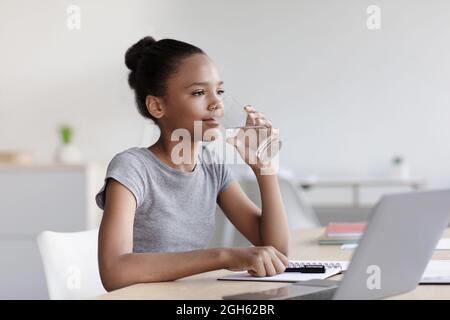 Une adolescente afro-américaine boit de l'eau du verre et étudie avec un ordinateur portable dans le salon Banque D'Images