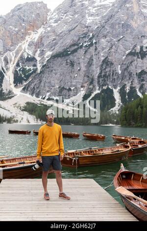 Un explorateur masculin en sweat-shirt jaune se tenant sur le fond du lac Braies avec des bateaux amarrés en bois dans les Dolomites Banque D'Images