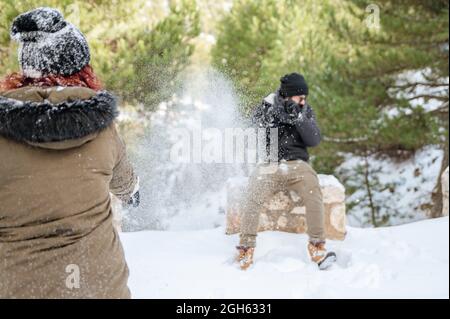 Des amis gais dans des vêtements chauds jouant des boules de neige dans la forêt tout en appréciant la journée d'hiver et en s'amusant Banque D'Images