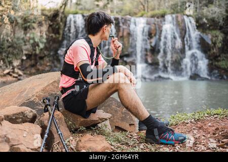 Vue latérale d'un randonneur mâle avec pack d'hydratation en train de boire au tuyau tout en ayant une pause pendant le trekking dans la forêt et en admirant la vue sur le lac près de la cascade Banque D'Images