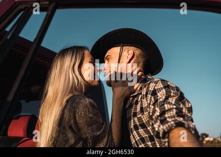 D'en dessous de la vue latérale de la jeune femme aimante embrassant homme dans le chapeau de cow-boy tendly près de la voiture sur fond de ciel bleu en soirée Banque D'Images