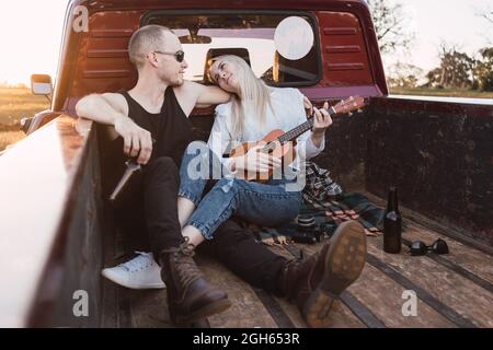 Couple aimant assis dans le coffre d'une voiture de pick-up d'époque et jouant ukulele tout en se refroidissant dans la campagne en soirée d'été et se regardant l'un l'autre Banque D'Images