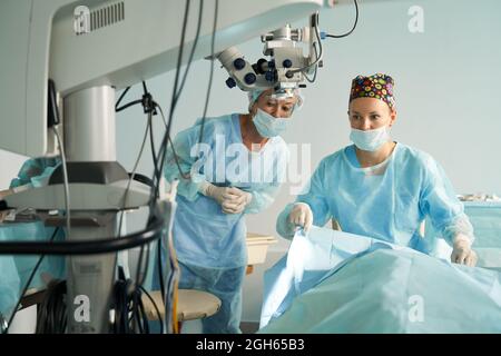 Femme médecin attentive en uniforme stérile contre un collègue qui se prépare à la chirurgie à l'hôpital avec un microscope Banque D'Images