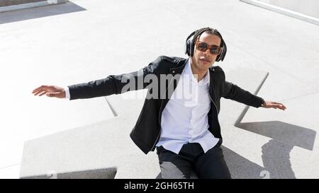 Grand angle de jeune homme ethnique avec des cheveux tressés vêtus d'un costume élégant et des lunettes de soleil pour écouter de la musique à l'aide d'un casque sans fil tout en se reposant sur l' Banque D'Images