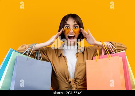 Wow, grande vente. Jeune femme émotive portant et touchant des lunettes de soleil tenant des sacs de shopping colorés, fond jaune Banque D'Images