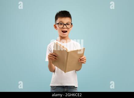 Un petit garçon asiatique enthousiaste portant des lunettes tenant et lisant un livre ouvert Banque D'Images