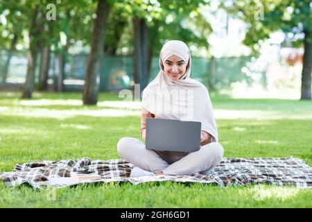 Joyeuse et attrayante femme islamique millénaire en hijab tapant sur un ordinateur portable, travailler à distance Banque D'Images