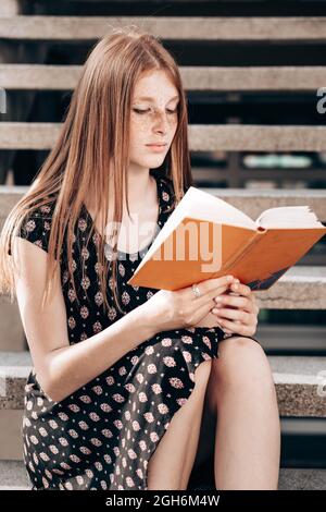 Jeune fille, gingembre, adolescente, assise sur les escaliers devant son école, lisant un livre. Banque D'Images