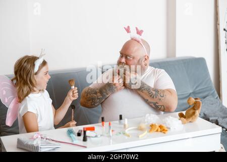 Un père effrayé avec serre-tête regarde une fille heureuse avec des brosses cosmétiques jouant au salon de beauté dans la chambre Banque D'Images