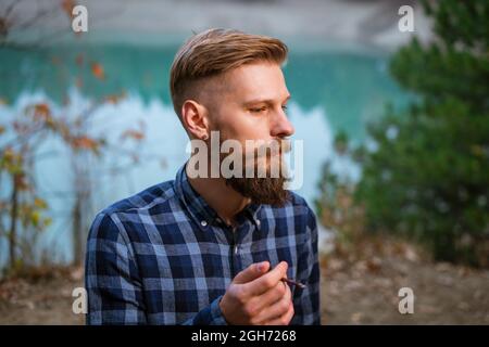 Fumer homme barbu à l'extérieur près du lac dans la soirée. Mauvaise habitude, dépendance à la cigarette. Un gars caucasien dans un dossier par le conducteur dans une chemise à carreaux fume une cigarette Banque D'Images