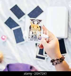 Main femelle avec clous violets tient la carte de tarot appelé le diable sur la surface blanche avec cahier ouvert et bougie. Vue de dessus. Minsk, Bélarus - 07.28.2021 Banque D'Images