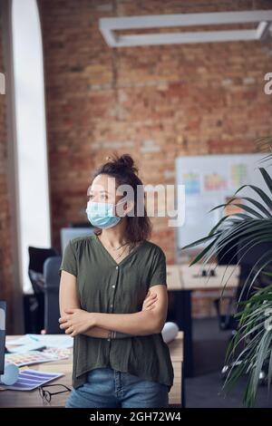 Femme d'affaires créative en tenue décontractée portant un masque de protection regardant de côté et se penchant sur la table tout en se tenant dans un bureau moderne vert sur un Banque D'Images