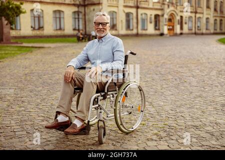 Prise de vue en longueur d'un patient handicapé âgé en fauteuil roulant regardant la caméra tout en passant du temps seul à l'extérieur près d'une clinique de réadaptation Banque D'Images