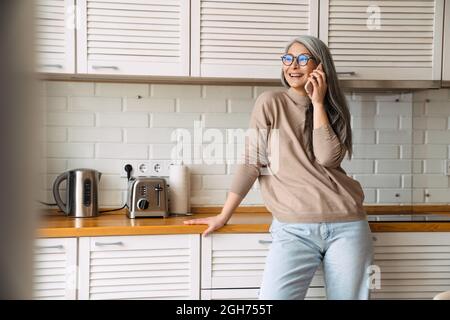 Femme mûre souriante parlant au téléphone portable tout en se tenant dans la cuisine à la maison Banque D'Images