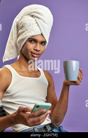 Un jeune homme dans une serviette sur la tête regarde le portrait de l'appareil photo. Souriant latino transgenre homme avec mobile Banque D'Images