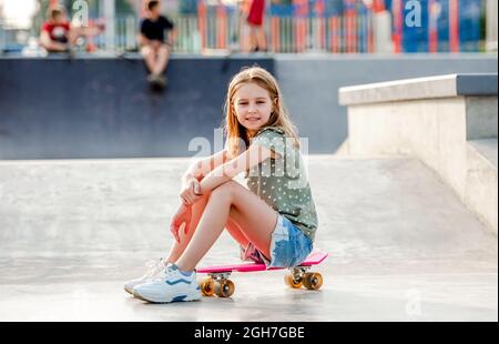 Préteen fille assise sur le skateboard à l'extérieur et regardant la caméra Femme patineuse enfant près de la rampe d'équitation de la ville en été Banque D'Images