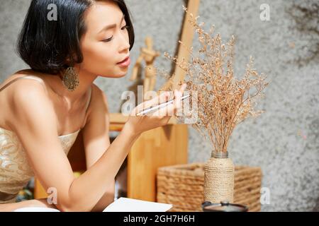 Belle jeune femme créative appréciant la beauté des fleurs rouges dans un vase sur une table basse Banque D'Images