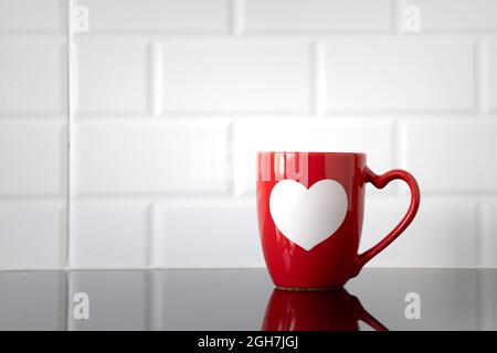 Une tasse à café rouge à coeur blanc sur une table de cuisine avec ses reflets sur la surface propre du comptoir. Banque D'Images