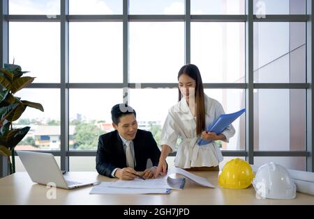 Le propriétaire du projet asiatique en costume et son assistant vérifiant le dessin de construction. Ambiance de travail matinale dans un bureau moderne. Banque D'Images