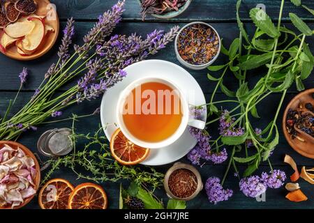 Thé. Herbes, fleurs et fruits autour d'une tasse de thé, un plat de dessus couché sur un fond rustique foncé de bois Banque D'Images