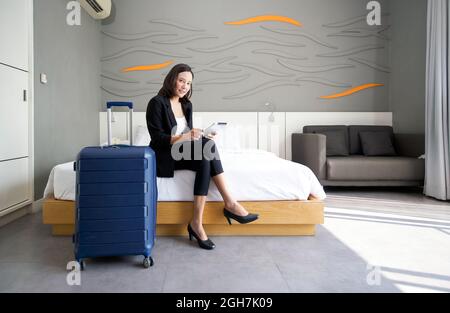 Femme d'affaires asiatique en costume noir assise avec un ordinateur tablette dans la main. Ses bagages ont été placés à côté du lit. Atmosphère de travail à l'extérieur pendant le Banque D'Images