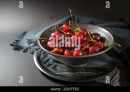 Cerises rouges fraîches dans un bol rustique en céramique avec serviette et assiette sur fond gris foncé, en-cas d'été sain, espace de copie, foyer sélectionné, étroit Banque D'Images