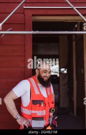 Homme barbu souriant ouvrier de construction debout avec la main sur la hanche à la porte Banque D'Images