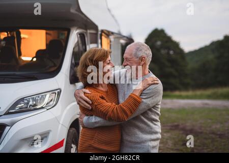 Couple senior debout et embrassant à l'extérieur au crépuscule, voyage de vacances en caravane. Banque D'Images