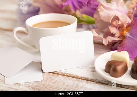 Carte de visite blanche avec une tasse de cioffee, bonbons au chocolat et fleurs d'iris sur fond de bois blanc. Vue latérale, espace copie, vie libre. Petit déjeuner, Banque D'Images