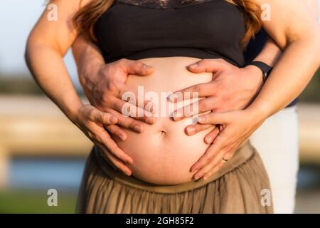 Vue rapprochée de quatre mains sur l'abdomen d'une femme enceinte adulte debout à l'extérieur Banque D'Images