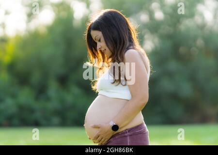 Femme enceinte touchant son abdomen dans un parc avec la lumière du soleil qui brille sur elle Banque D'Images