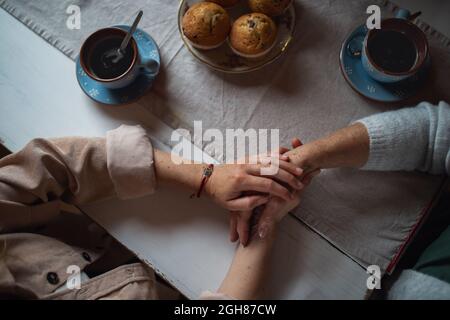 Vue de dessus de la mère senior ayant un café avec une fille adulte à l'intérieur à la maison, tenant les mains. Banque D'Images