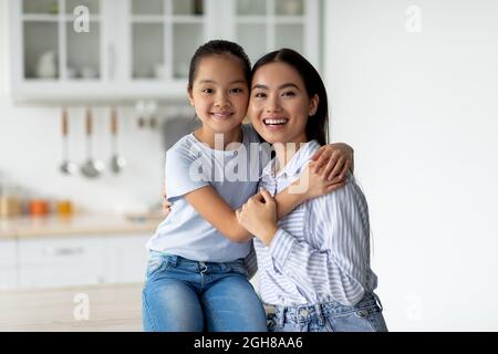 Portrait de la jeune mère asiatique et de la petite fille mignonne posant dans la cuisine intérieure, jolie fille embrassant sa mère Banque D'Images