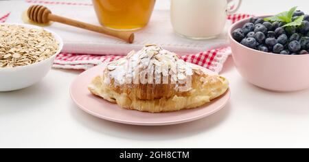 croissant cuit arrosé de sucre en poudre, de bleuets et de flocons d'avoine dans une plaque en céramique sur une table blanche, petit déjeuner Banque D'Images