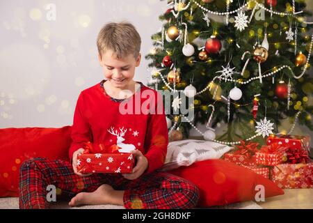 Portrait de garçon excité en pyjama rouge tenant un cadeau de Noël. Émotions positives pendant les vacances de Noël. Banque D'Images