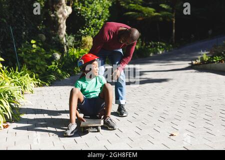 Père afro-américain avec son fils souriant et jouant avec le skateboard dans le jardin Banque D'Images