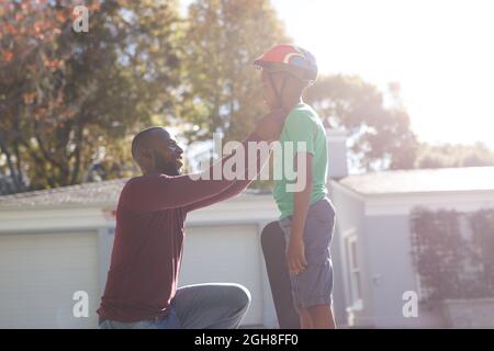 Père afro-américain avec son fils souriant et se préparant avant de skate dans un jardin ensoleillé Banque D'Images