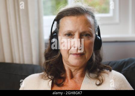 Portrait d'une femme caucasienne âgée heureuse dans le salon, assise sur un canapé, portant un casque Banque D'Images