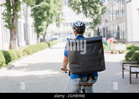 Vue arrière du coursier dans un casque et sac à dos thermo à vélo dans la rue urbaine Banque D'Images