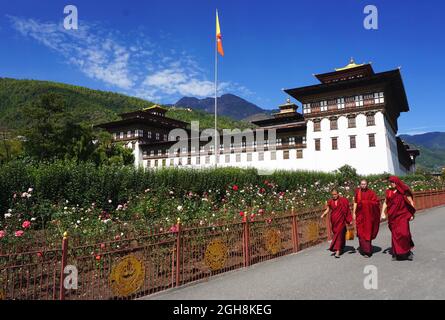 Trois moines bouddhistes aux robdes rouges se promeissent devant de magnifiques jardins fleuris devant Tashichho Dzong, le siège officiel du gouvernement bhoutanais depuis 1952. Banque D'Images