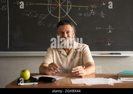 Professeur avec visage amical regardant la caméra assis à son bureau dans la salle de classe avec le tableau noir dans l'arrière-plan. Banque D'Images