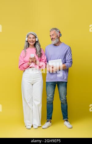 couple d'âge moyen, multiethnique et souriant, dans un casque sans fil avec gadgets sur fond jaune Banque D'Images