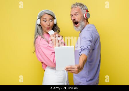 j'ai surpris un couple d'âge moyen et interracial dans un casque sans fil en regardant une tablette numérique isolée sur jaune Banque D'Images