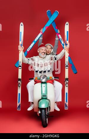 heureux skieurs interraciaux et d'âge moyen dans des chandails tenant des skis tout en faisant du scooter de moteur sur rouge Banque D'Images